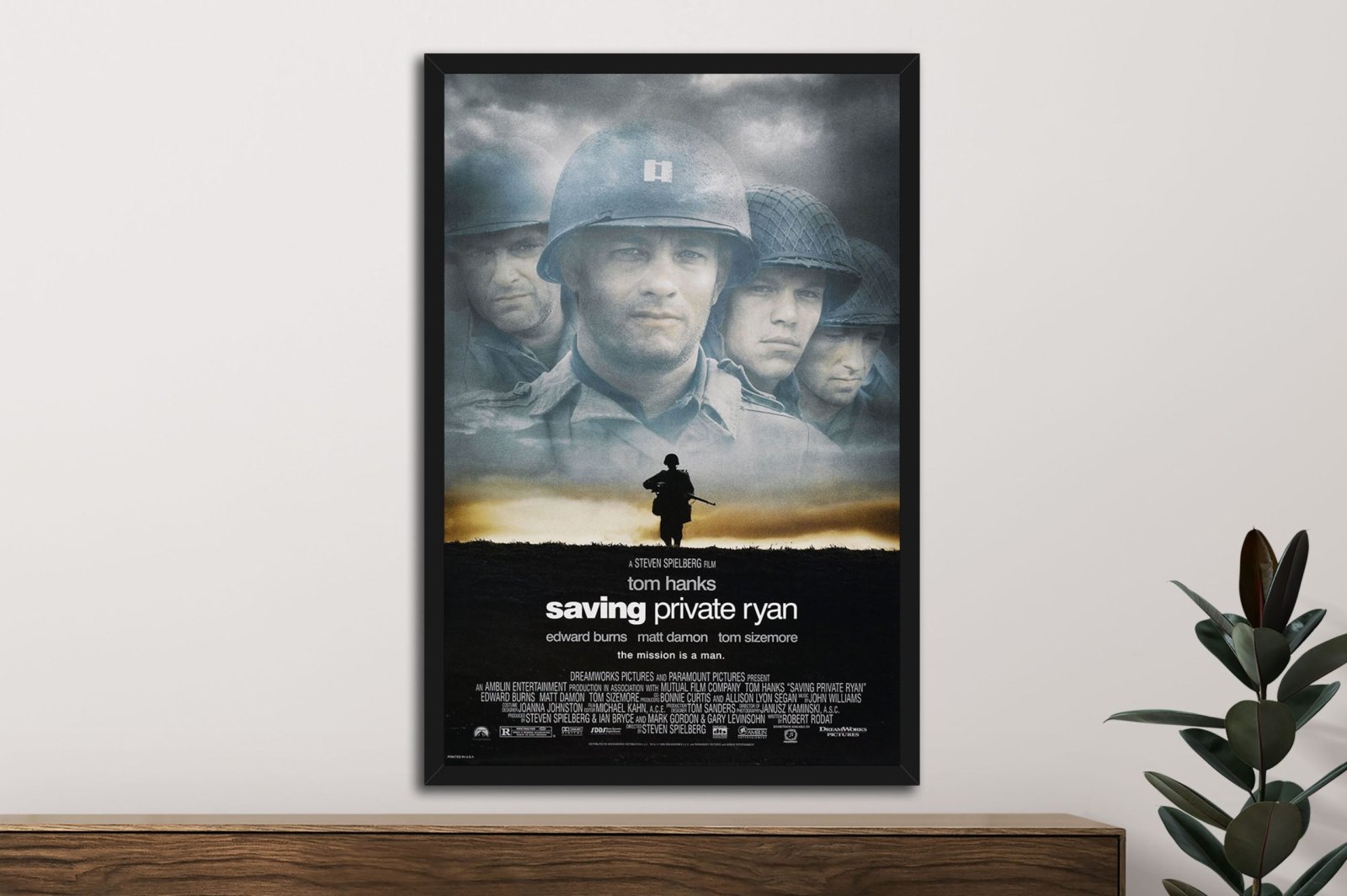 Framed Saving Private Ryan movie poster on white wall above timber sideboard with rubber plant, showing Tom Hanks and soldiers