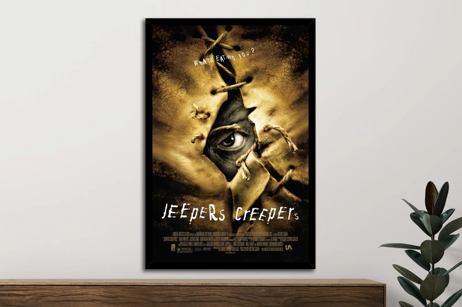 Framed Jeepers Creepers movie poster on white wall above timber sideboard, dark sepia artwork with stitched eye