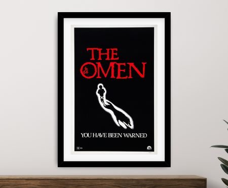 The Omen vintage horror film poster framed in black, featuring red title and eerie child silhouette on a neutral wall
